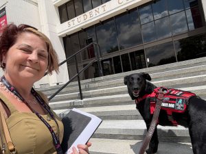 risha Silvasy stands beside Loki, her half Border Collie, half Labrador companion. Loki, a large black dog is wearing a red and gray service harness.