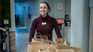 Campus Dietitian and Food Service Manager Jessica Carr, wearing a maroon turtleneck and name badge, stands behind a counter with several brown paper Farm Box bags ready for pickup. The setting is a campus dining facility.