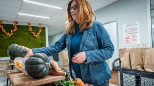 Jennifer Bostedo, wearing a blue jacket and standing indoors, arranges dark green acorn squash and other fresh produce on a wooden table as part of a Farm Box display.