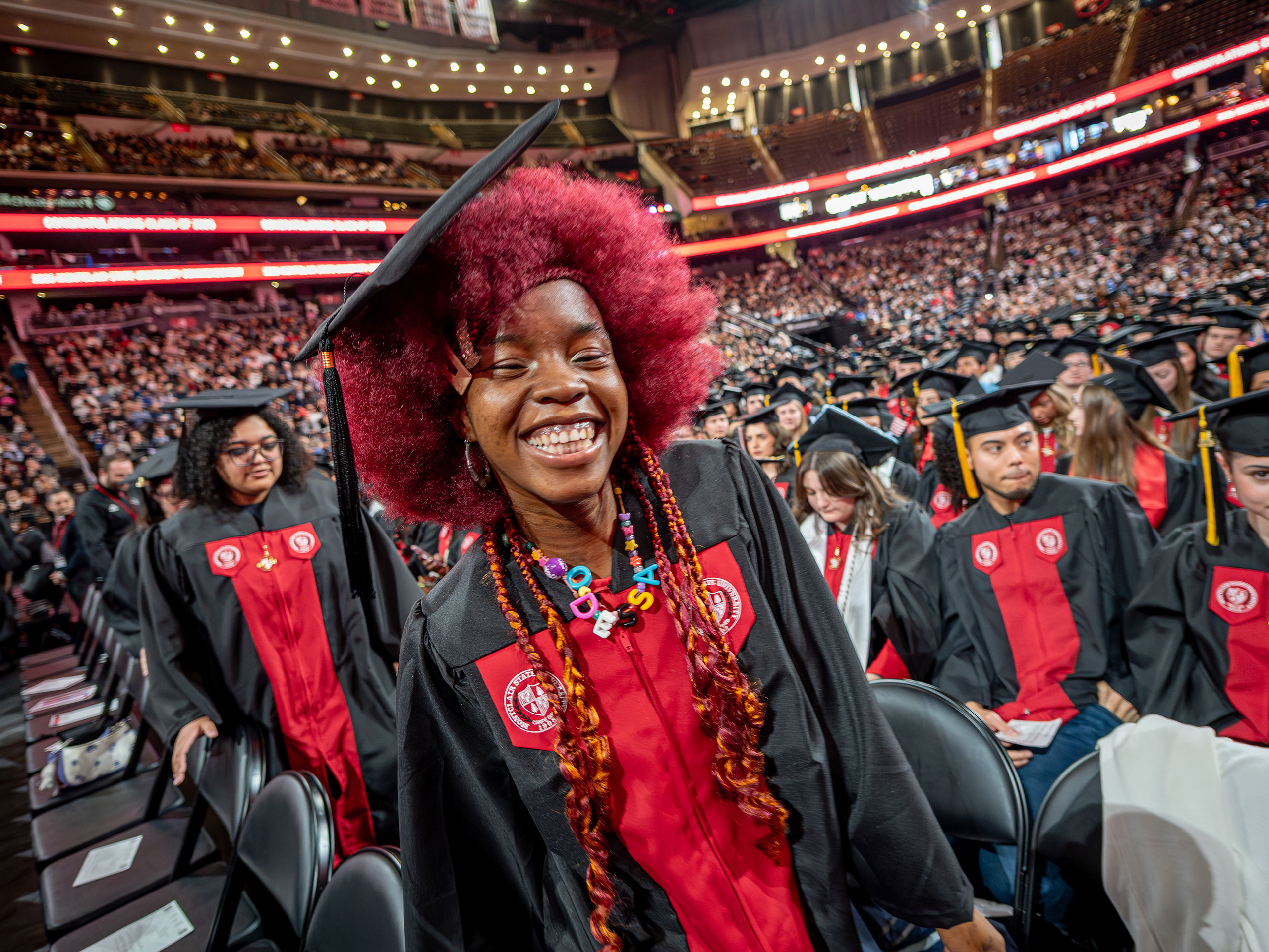 A smiling undergraduate in a black gown and cap walks between rows of seated classmates, her bright red hair and colorful beaded necklace standing out.