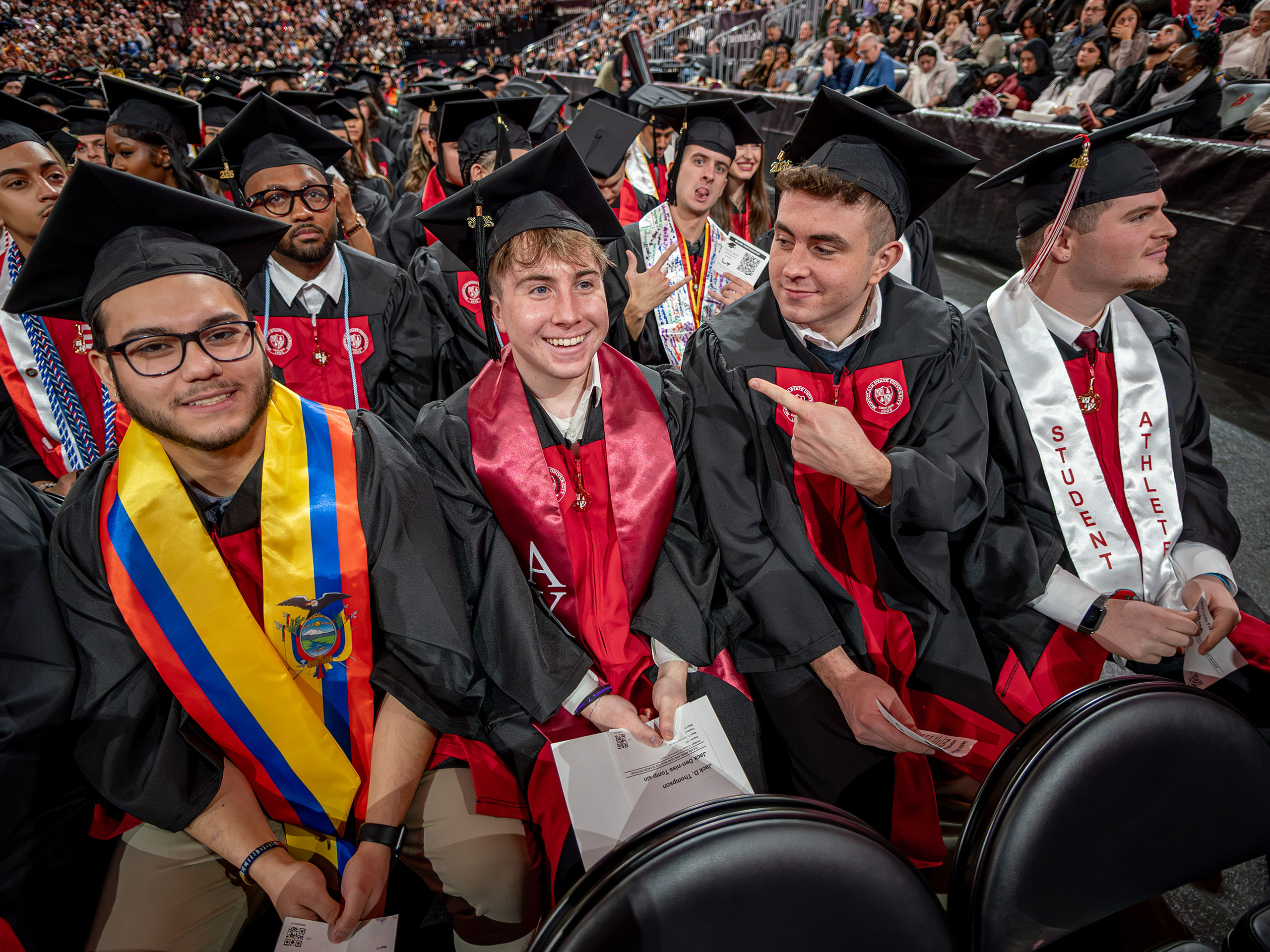 Four undergraduate students in black gowns and caps sit together in a row, smiling and laughing, with one student playfully pointing at a classmate.