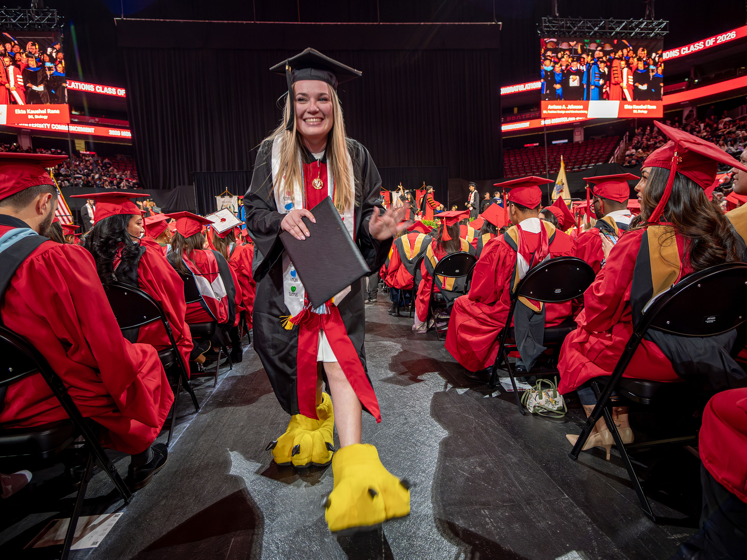 An undergraduate in a black gown and cap walks down the aisle holding her diploma cover while wearing oversized yellow mascot feet, continuing the tradition in which students who served as the college mascot reveal themselves at graduation.