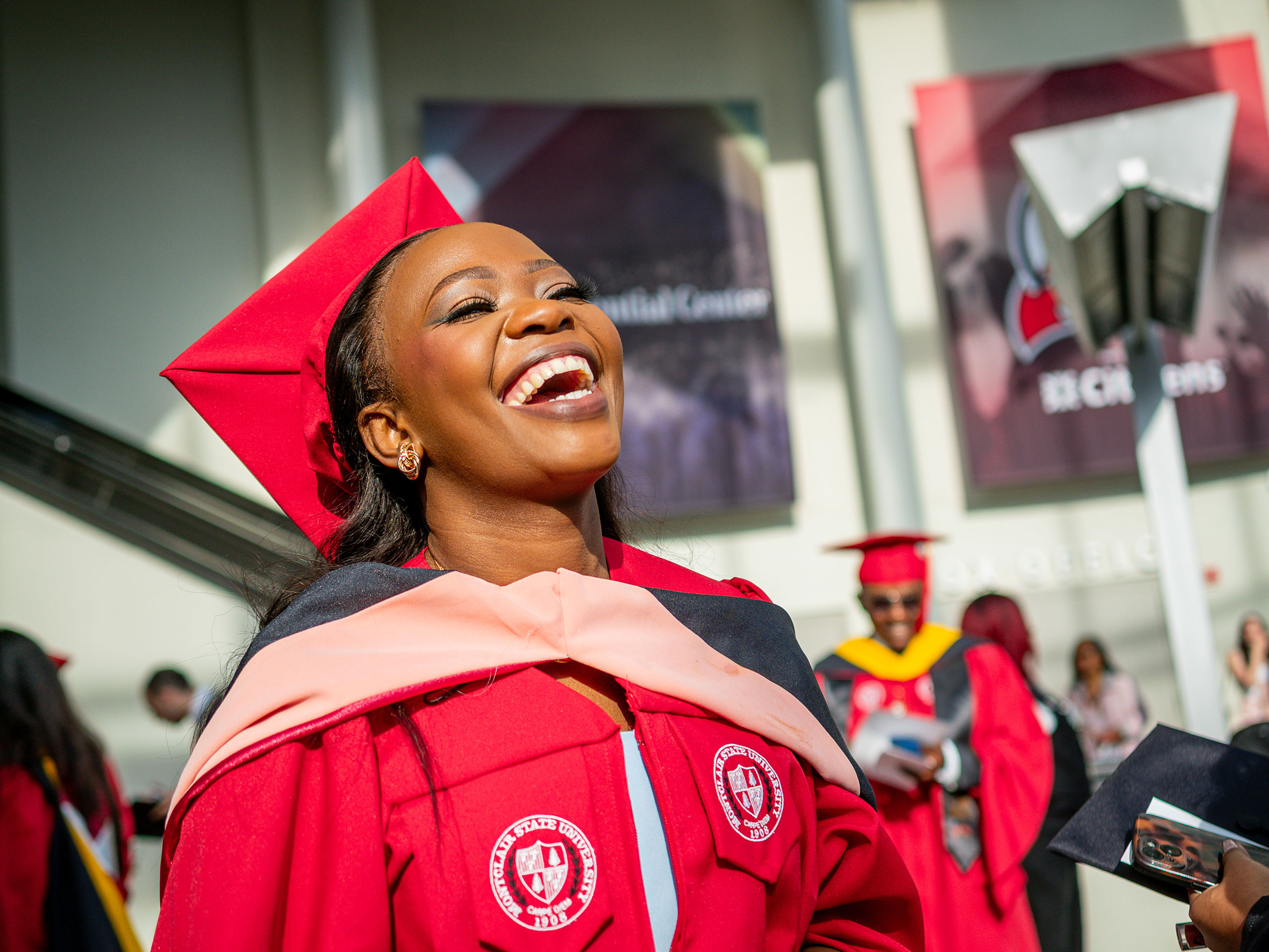 A Montclair State University Graduate School student in a red cap and gown smiles and looks upward in the bright lobby of Prudential Center.