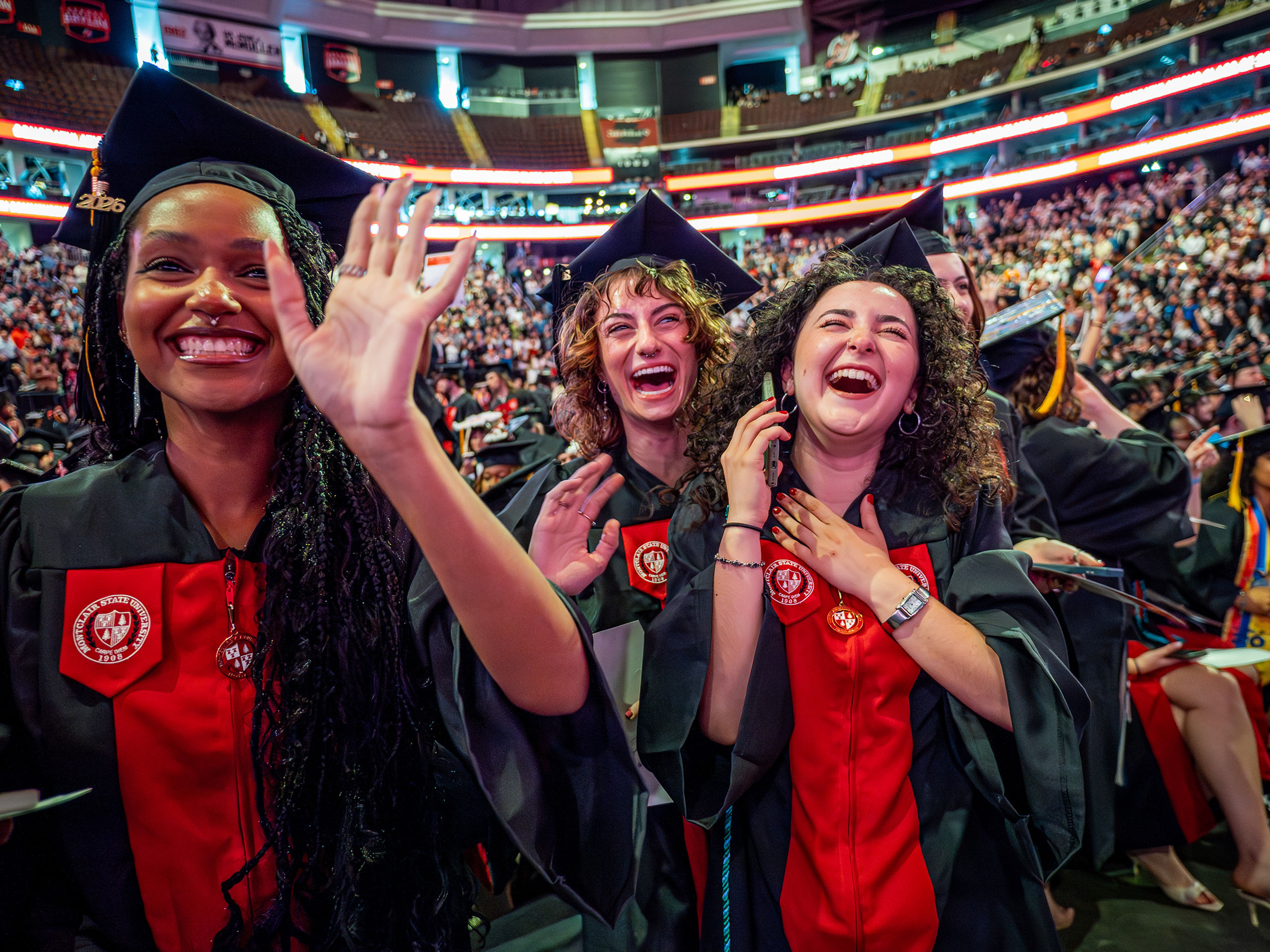 Three Montclair State University graduates in caps and gowns smile, laugh and wave from their seats on the arena floor, surrounded by rows of fellow graduates and guests during the 2026 Spring Commencement ceremony.