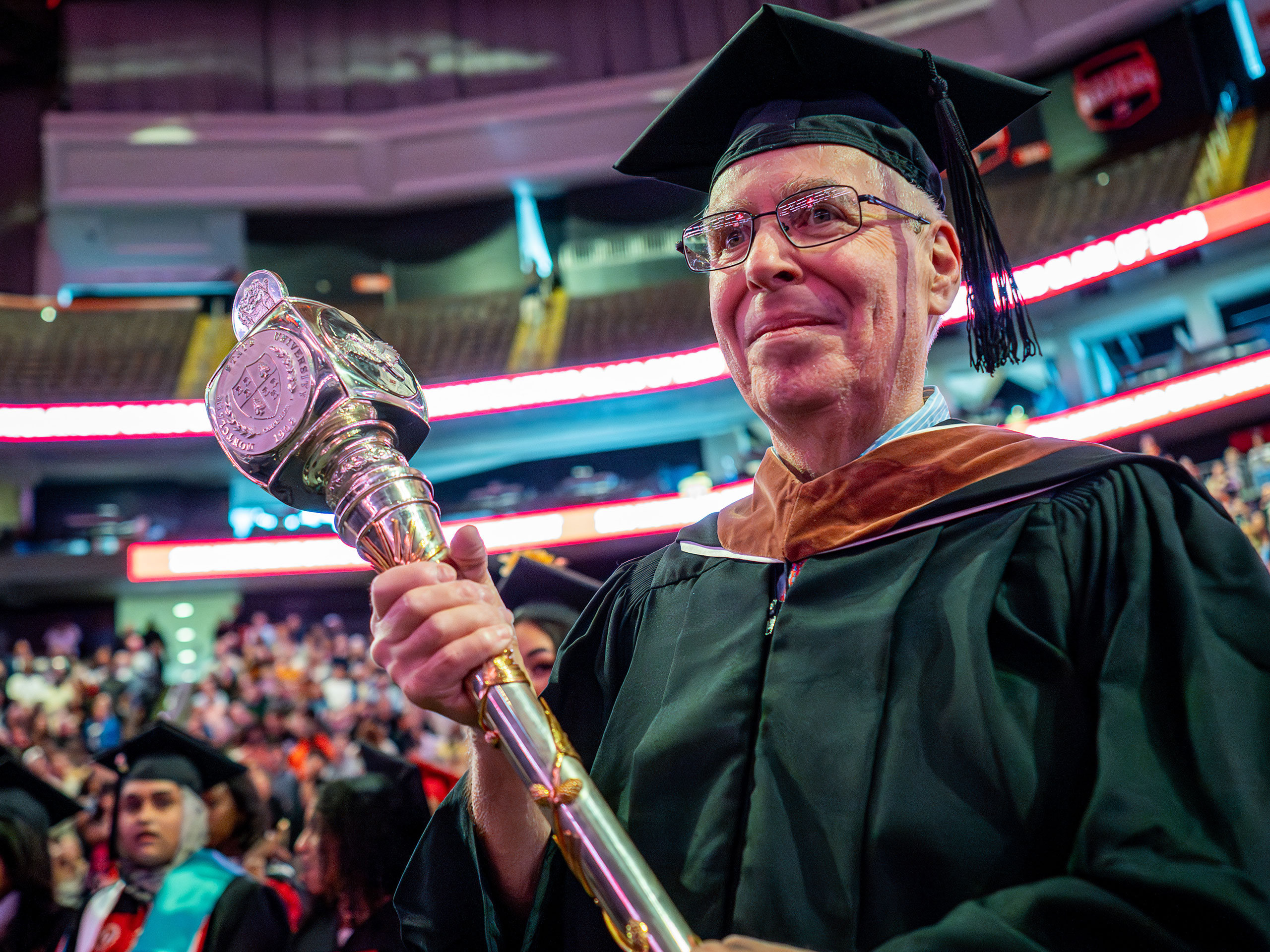 Grand Marshal Eric Diamond, a Montclair State University Theatre and Dance professor, wears academic regalia and holds the ornate silver University mace while leading the 2026 Spring Commencement procession inside Prudential Center.
