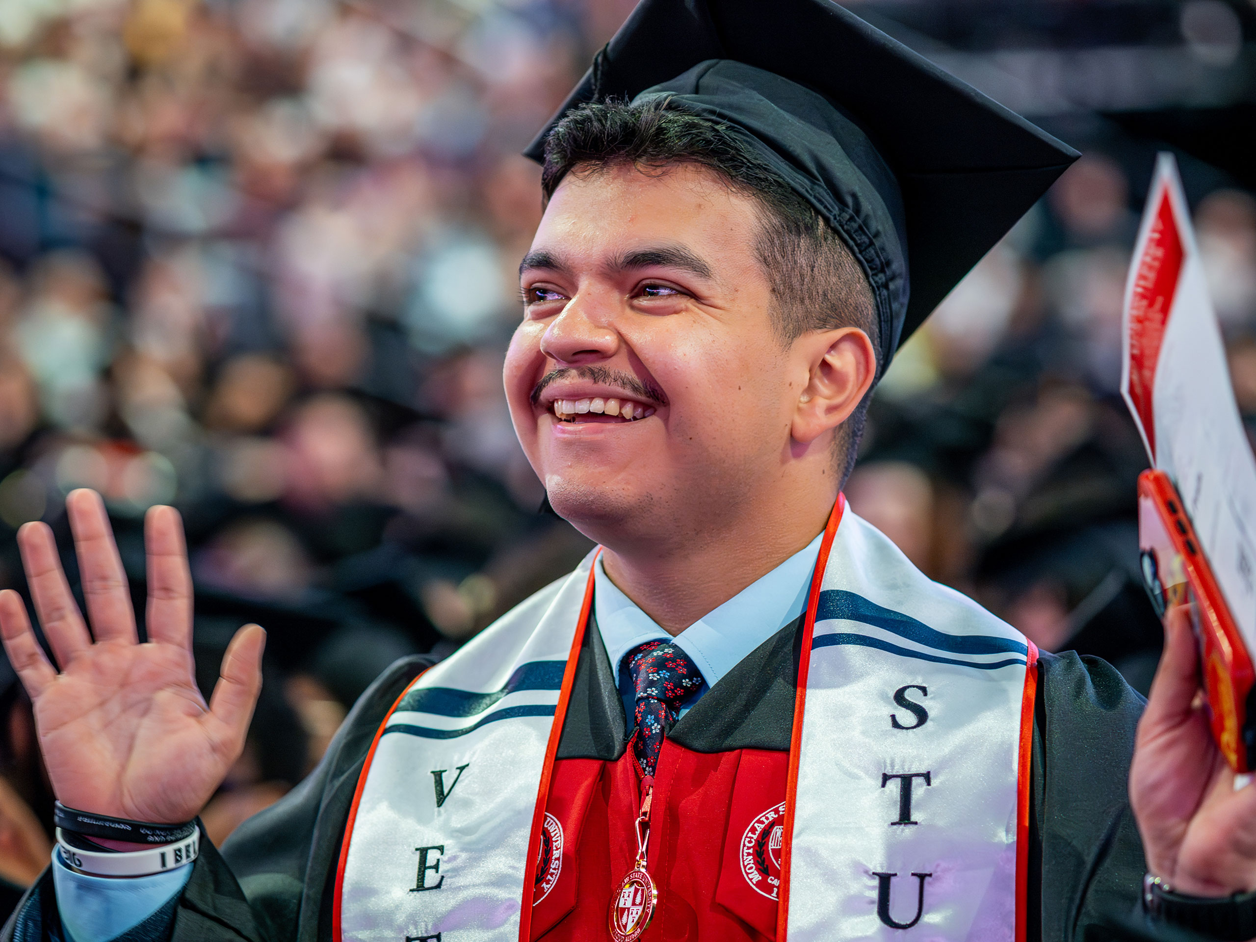 A smiling Montclair State University graduate in cap and gown raises one hand in celebration during 2026 Spring Commencement, wearing an honor stole and standing before a blurred crowd of fellow graduates.