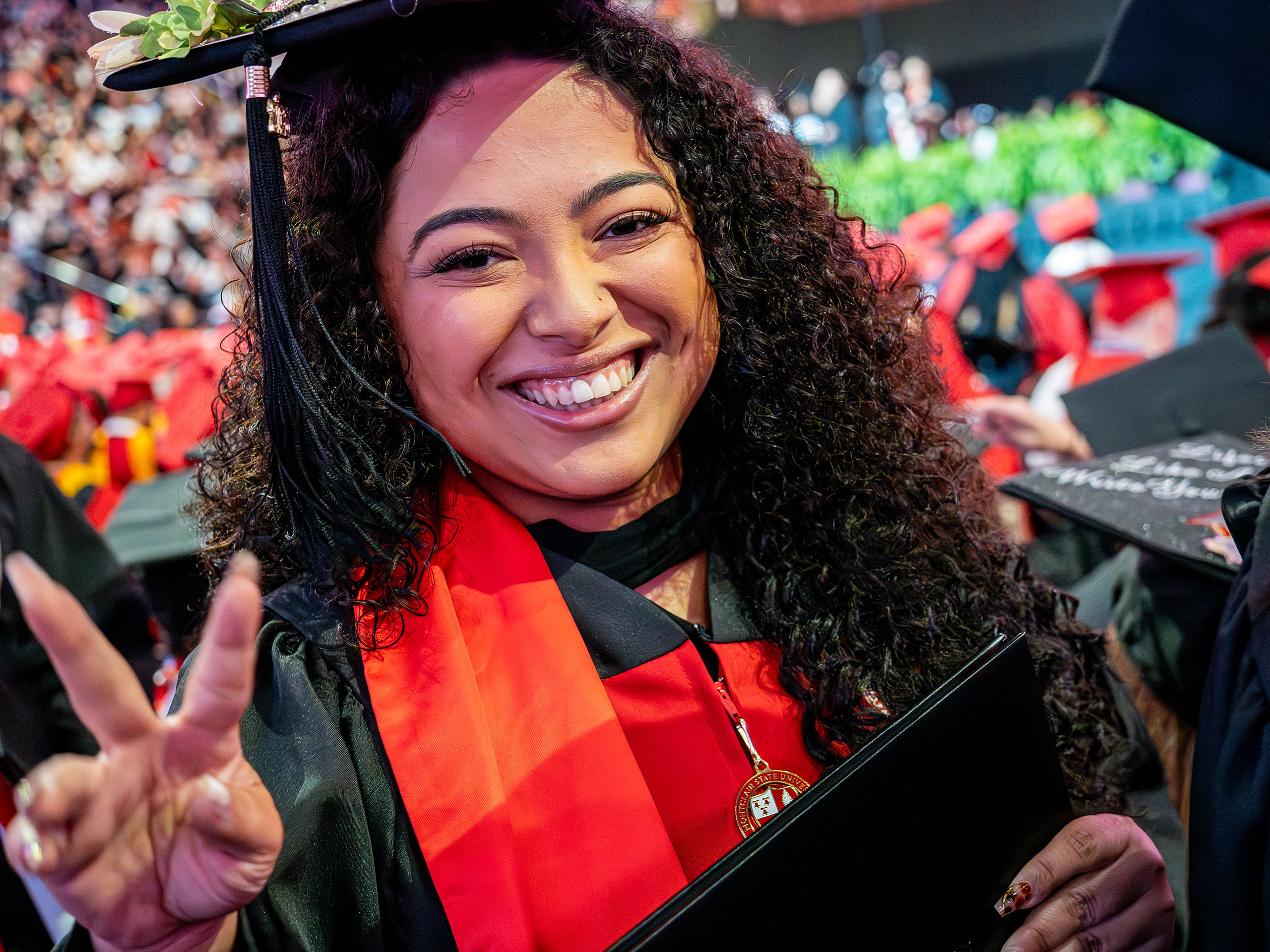 A Montclair State University graduate in black cap and gown smiles broadly, holding a diploma folder and flashing a peace sign during the 2026 Spring Commencement ceremony.