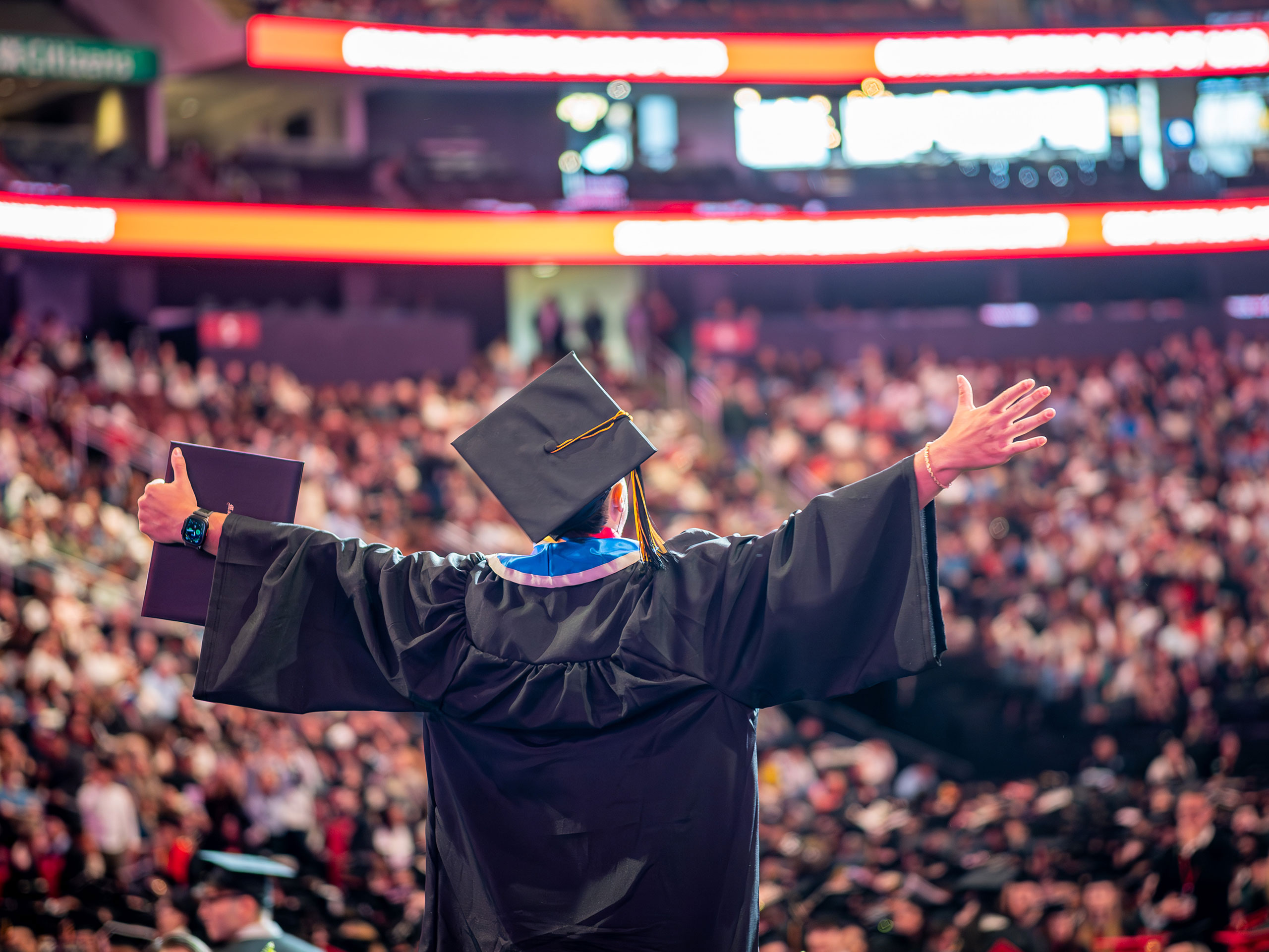 A graduate in a black cap and gown stands on stage with arms outstretched and diploma in one hand, facing an arena filled with cheering guests during the 2026 Spring Commencement ceremony.