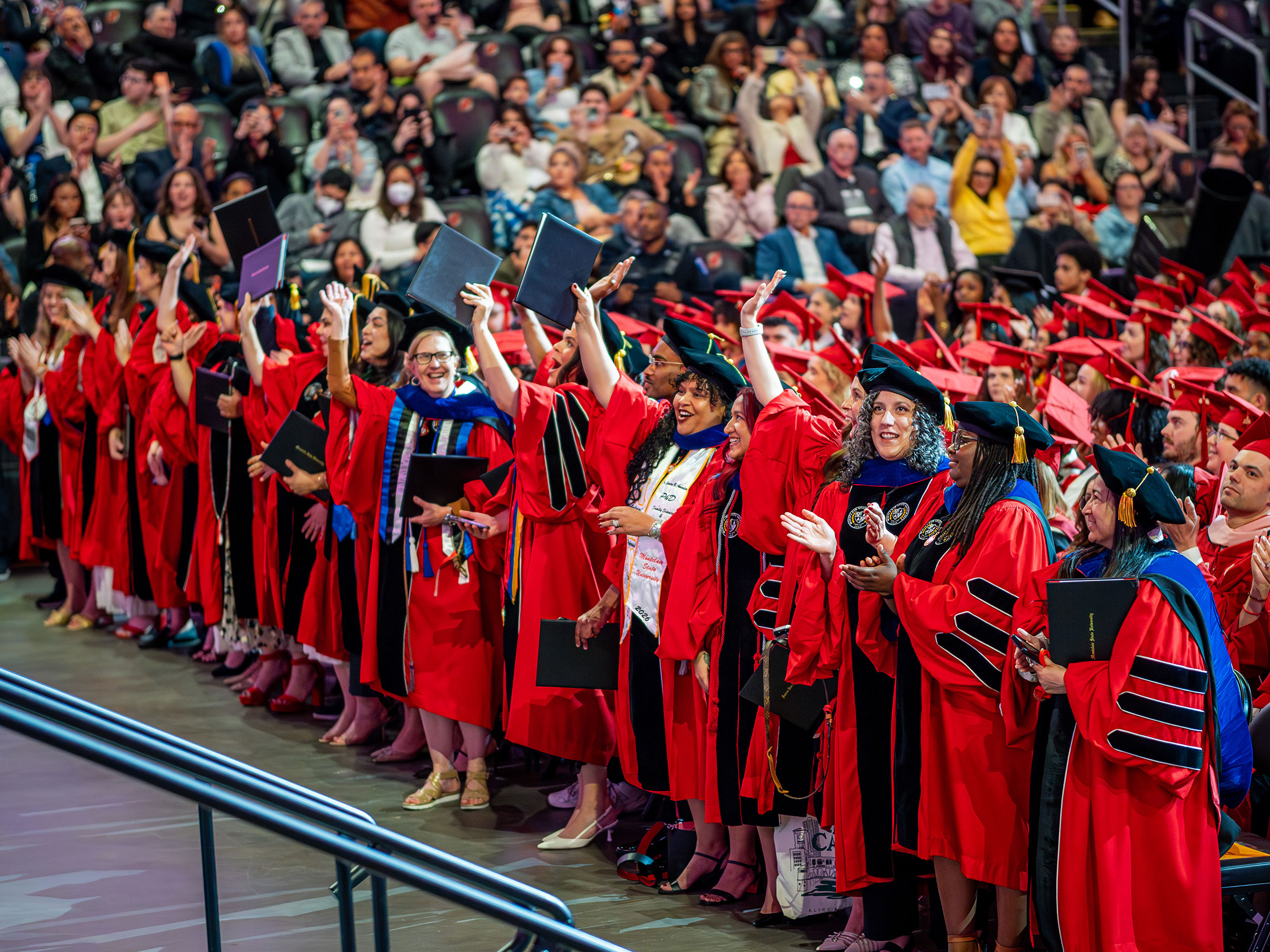 A row of Montclair State University students who have earned doctoral degrees stand in bright red academic regalia, holding diplomas and raising their hands in celebration as the audience in the arena seats applauds behind them during the 2026 Spring Commencement ceremony.