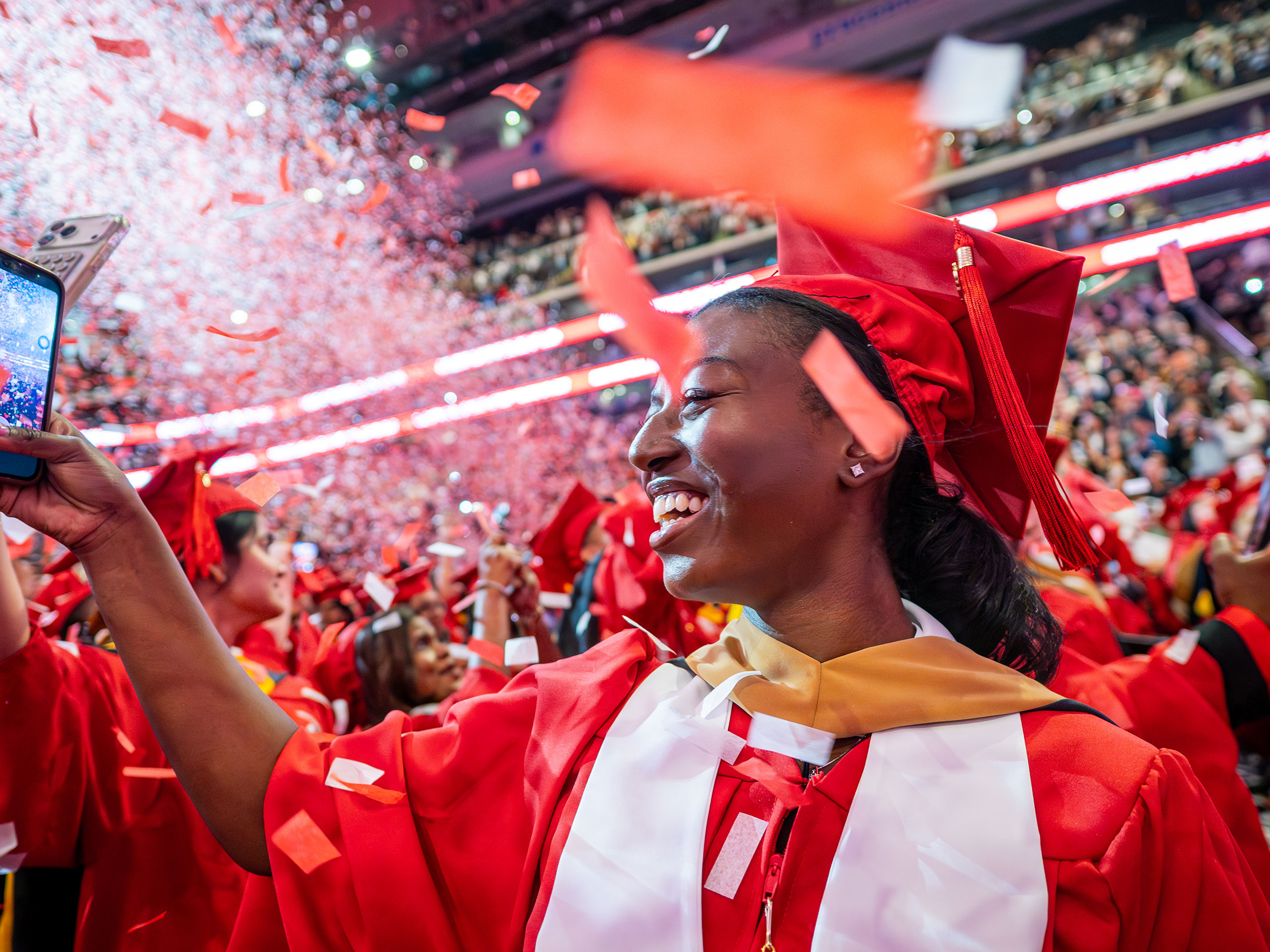 A Montclair State University graduate in a red cap and gown smiles and holds up a phone to capture the moment as red and white confetti rains down around her and fellow graduates during the 2026 Spring Commencement ceremony.
