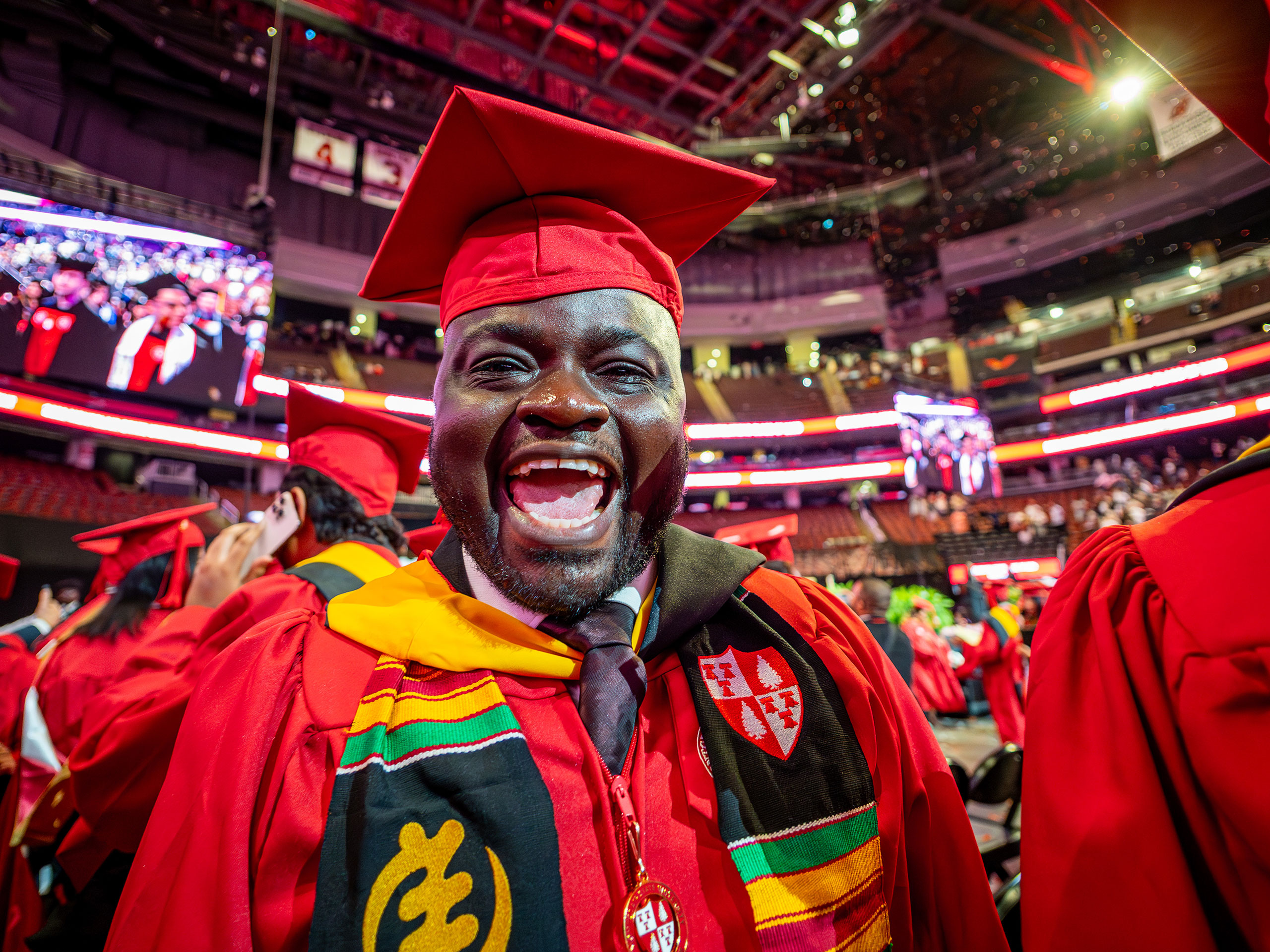 A Montclair State University graduate in a red cap and gown smiles broadly at the camera, wearing a colorful kente stole and standing among fellow graduates on the arena floor during the 2026 Spring Commencement ceremony.