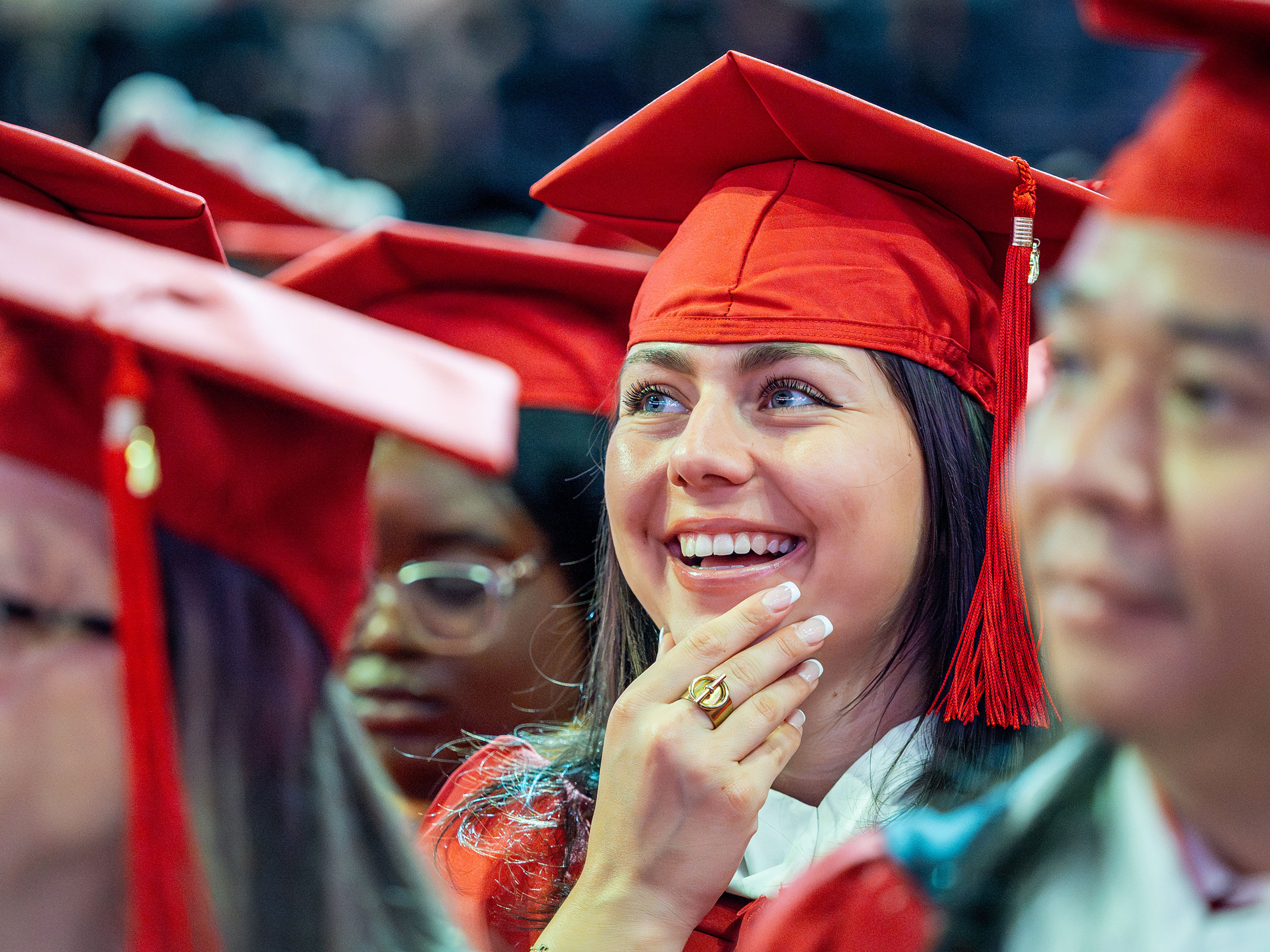 Graduate students wearing red caps and gowns sit together during Montclair State University’s 2026 Spring Commencement, with one graduate in the foreground resting a hand near their face as they listen to the ceremony.