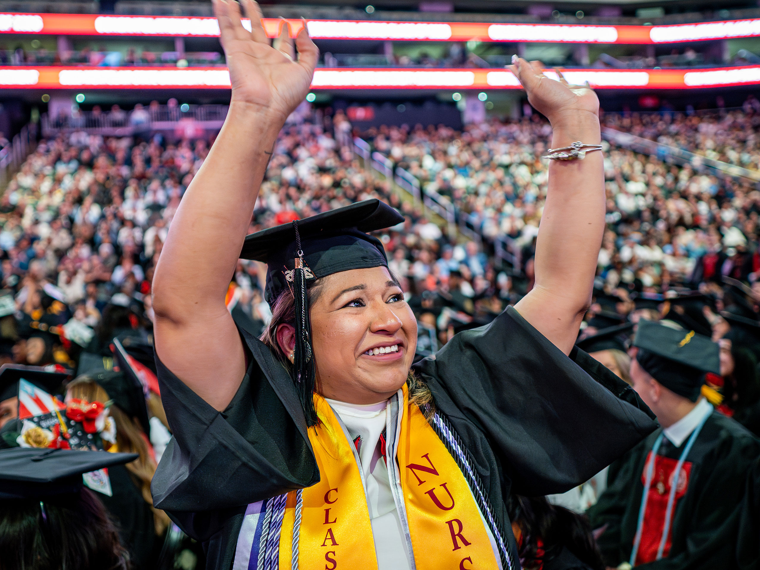Nursing graduate in cap and gown raises both arms in celebration during Montclair State University’s 2026 Spring Commencement, wearing a yellow “Nursing” stole that signifies her program, with a full arena audience in the background.