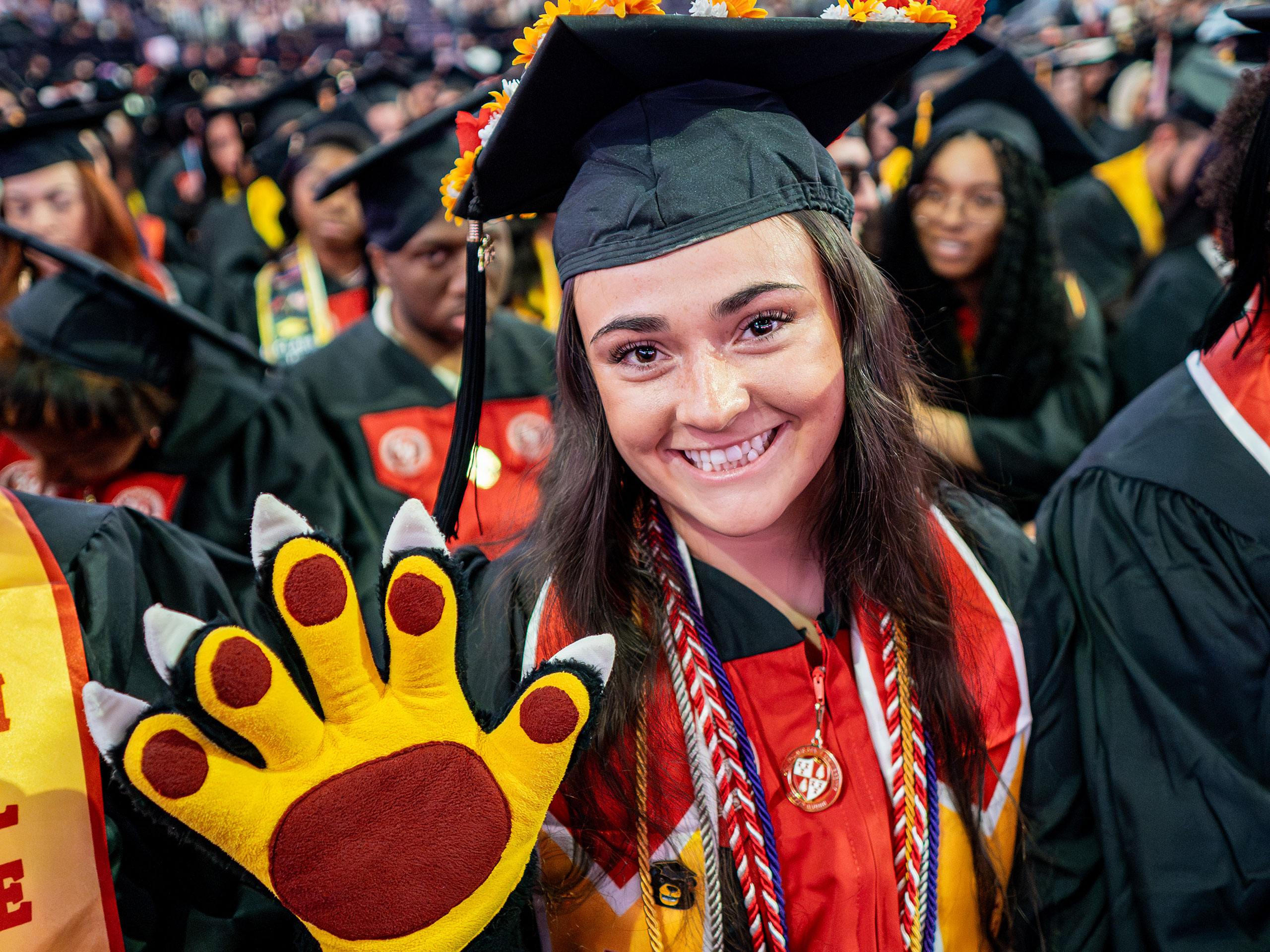 Graduate in cap and gown holds up a large yellow bear-claw mascot glove during Montclair State University’s 2026 Spring Commencement, continuing the Bloomfield tradition of revealing mascot identities at graduation.