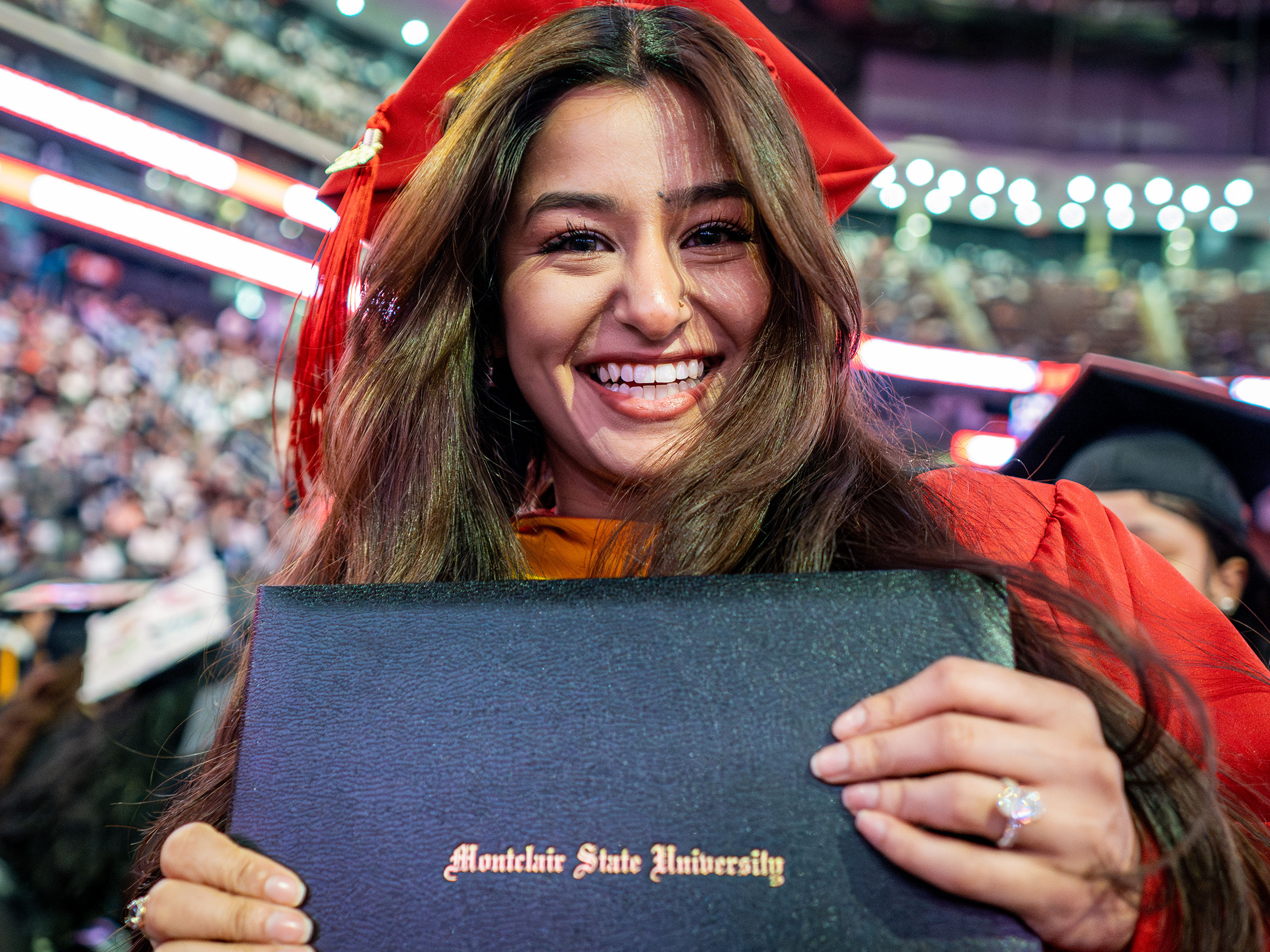 Graduate in red cap and gown holds up a Montclair State University diploma cover toward the camera during the 2026 Spring Commencement ceremony, with the arena crowd blurred in the background.