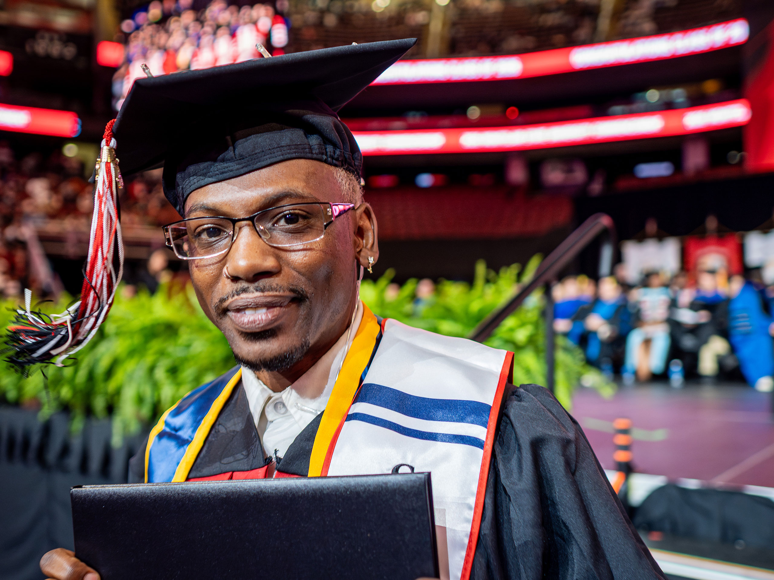 Graduate in cap and gown holding a diploma walks past the stage at Montclair State University’s 2026 Spring Commencement, with faculty seated and bright arena screens visible in the background.