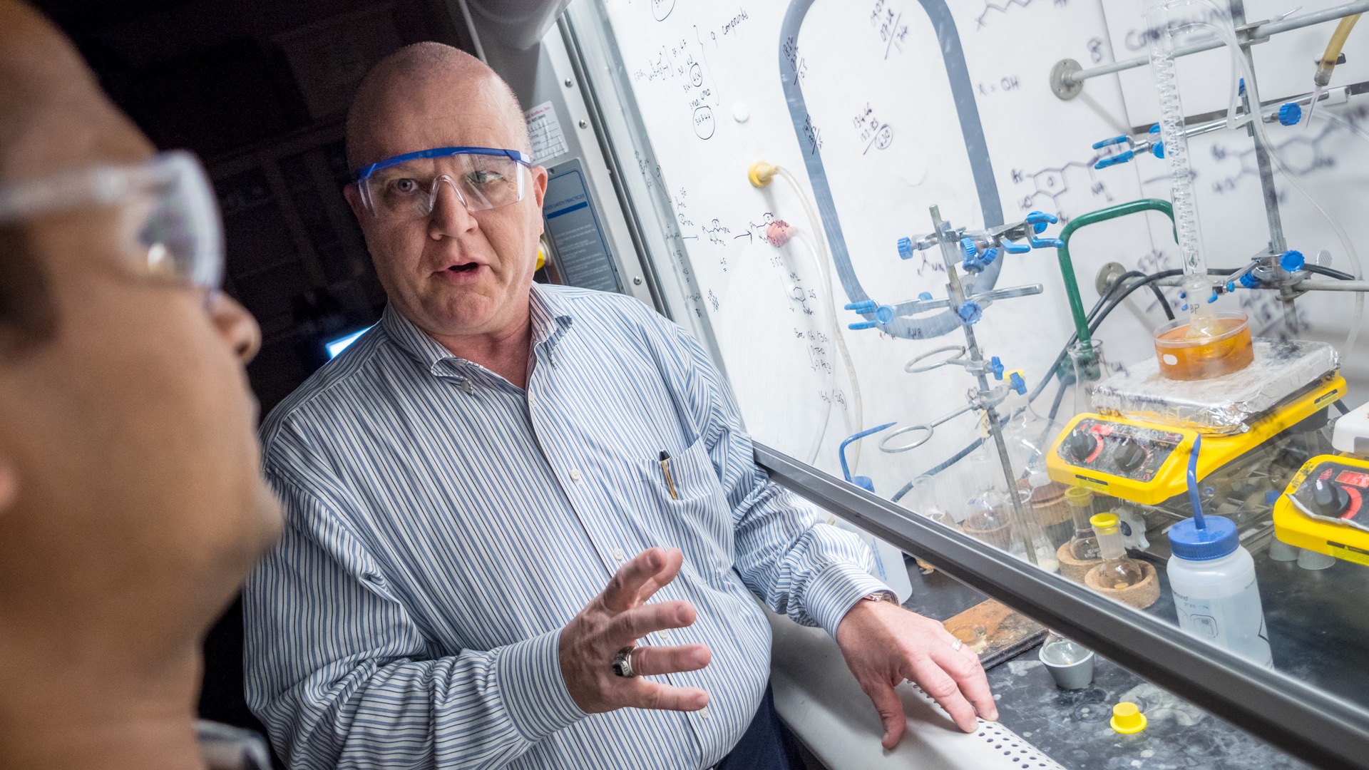 Two university staff wearing safety goggles and standing near a lab hood with chemistry equipment.