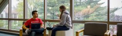 Two students sitting with laptops and talking in front of windows