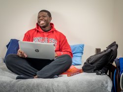 Photo of student using laptop while sitting on bed in residence hall.