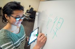 A student stands at a whiteboard in a classroom, writing Chinese characters.