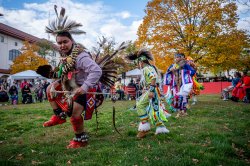 A group of Indigenous dancers dressed in traditional regalia adorned with feathers, beads and intricate patterns, perform outdoors on a grassy area during a cultural event.