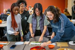 A group of students stands around a table making sushi using bamboo mats and hands, picking up fillings with chopsticks. Various sushi ingredients such as cucumber and carrot sticks, egg slices as well as bowls and plates are arranged on the table. The activity takes place in a classroom with others participating in the background.
