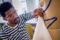 Student wearing glasses and a striped shirt measuring a piece of fabric against manikin