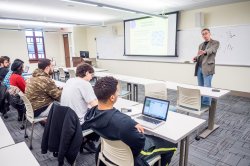 Professor speaking to students in a small classroom with projection screens behind