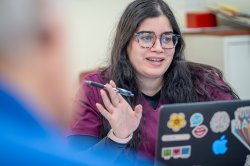 A woman with pen in hand converses with her colleagues while seated behind a laptop adorned with stickers that have messages of positivity.
