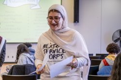 Student wearing hijab and a t-shirt that reads "next gen service corps" holding papers and speaking to another person