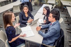 four students sit at a table with notebooks and laptops open between them