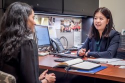 A woman behind a desk in an office speaks with a peer sitting across the desk.