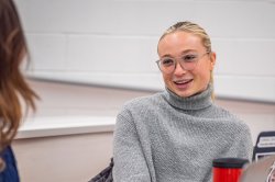 student in grey sweater and glasses speaking with someone in the foreground wearing a blue dress