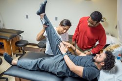 One student laying on a table describes how he's feeling to a trainer as the trainer stretches the student's hamstring. Another student looks on, observing the trainer.