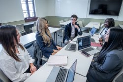 Five students having a discussion sit at a classroom table with open laptops.