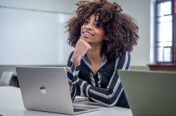 woman student with laptop sits at table in classroom resting her chin on her hand and smiling