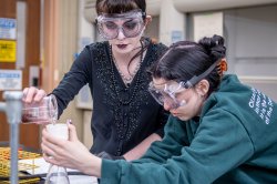 One student pours a liquid solution from a beaker while another student holds a funnel atop an Erlenmeyer flask
