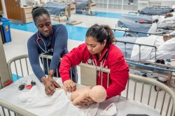 Two nursing students in a hospital-like classroom reach into a crib to practice procedures on a baby test dummy