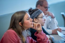 Three students sit in a classroom; two are listening and one is taking notes.