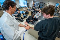 A teacher holds a magnetic coil designed to detect neural activity over a student's arm in front of a lab full of students