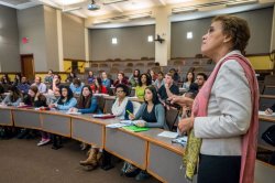 A college professor stands at the front of a lecture hall, addressing a classroom filled with students who are seated in curved rows and taking notes.