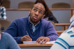 A student takes notes in a lecture hall with other students in the foreground and background.