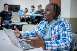 In a classroom, a student with an open laptop gestures while speaking.