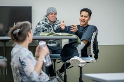 A student seated at a high table with barstools speaks while gesturing with his hands, his professor out of focus in the foreground listening.