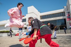 A student dressed in a pink martial arts uniform performs a high jump kick during a Taekwondo workshop held outdoors in front of the Student Center, while an instructor in a black uniform holds a target pad.