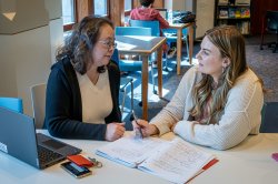A professor and a student meet in a lounge area with a laptop and a notebook full of notes open on the table.