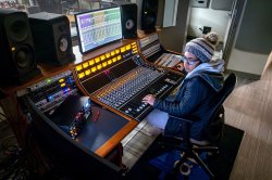 A student sits in front of a large mixing board in a professional-grade recording studio, the performance space visible through a window in front of her.