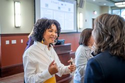 A student has a dialogue with a professor in a large meeting area with her sales pitch slides displayed on the large screen behind her.