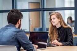 Two students sit across from each other with laptops open on the table in between them