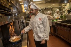 A student in a white chef's jacket and hat cooks food in a frying pan in a combination laboratory and kitchen